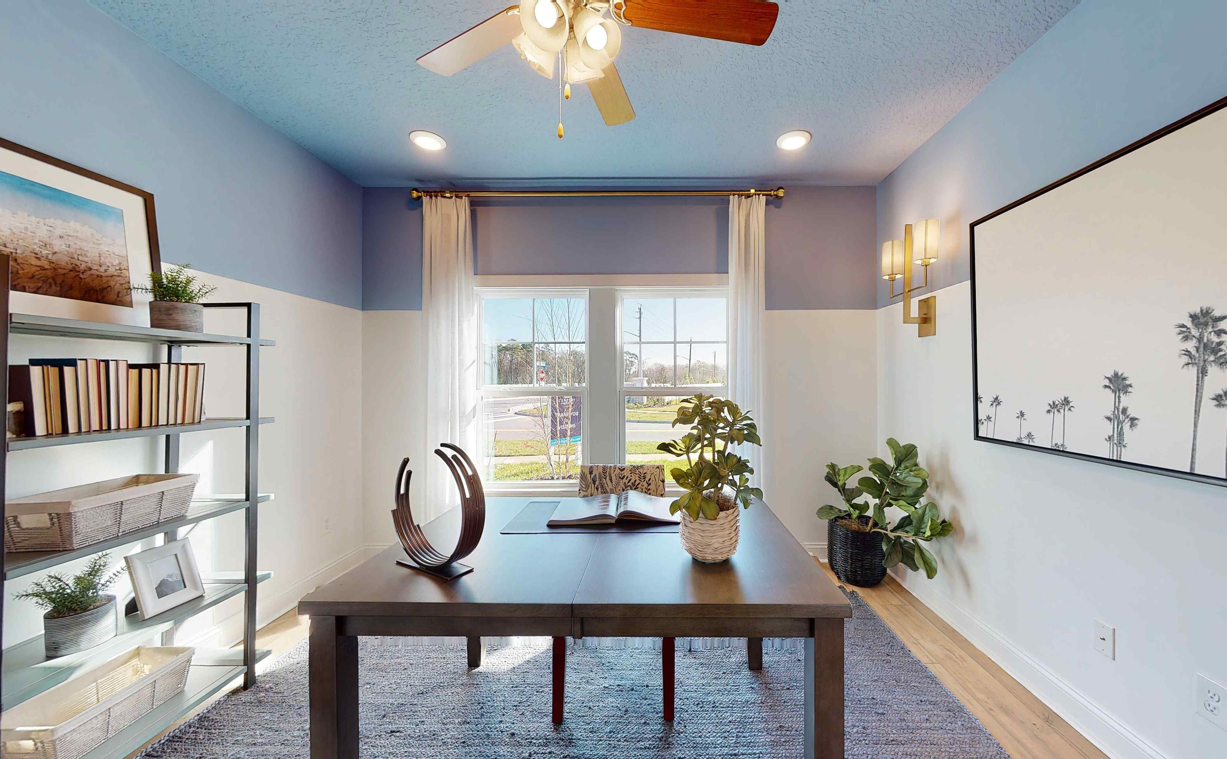 A room with a table and a window with a fan and a plant.